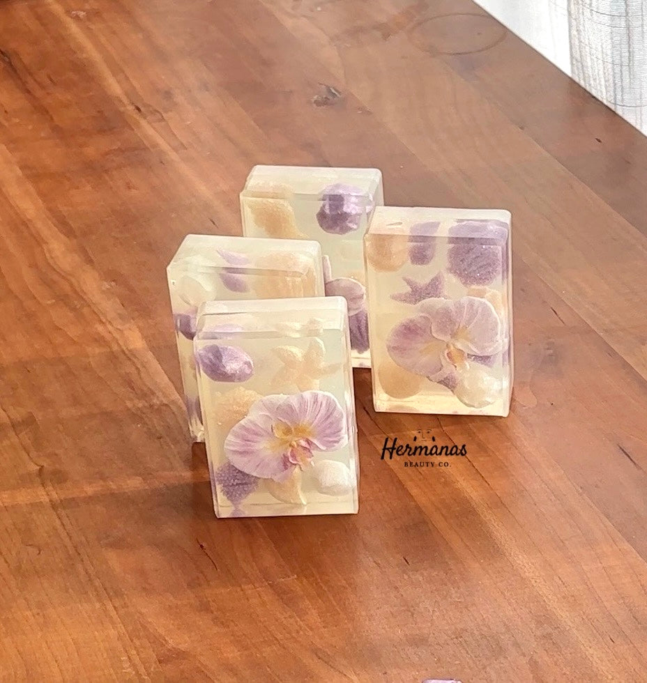Three square soap bars with floral designs on a wooden surface, branded 'Hermans Beauty Co'.