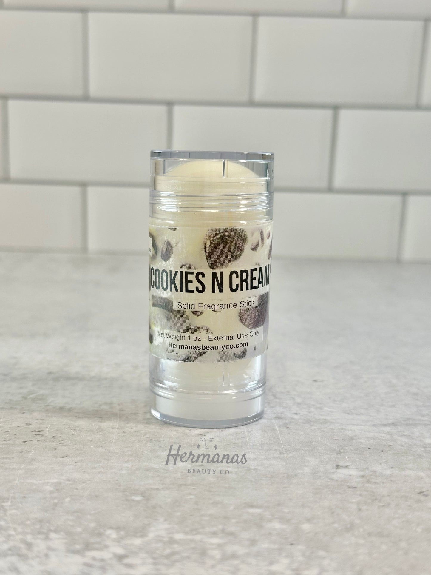 Cookies N Cream solid fragrance stick in a clear container on a light gray surface with a tiled wall background.