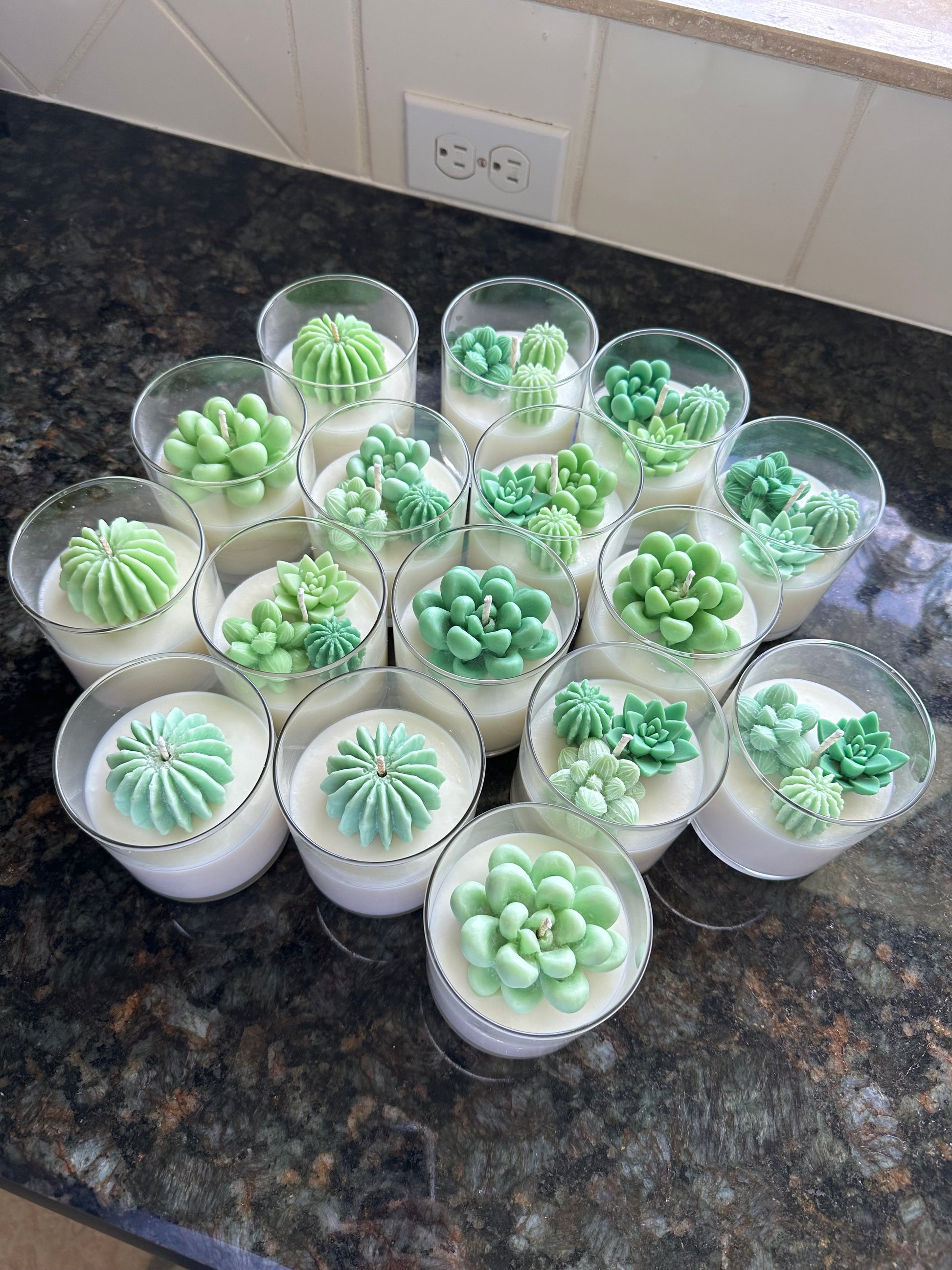 The image shows a group of candles arranged in a close-knit cluster on a dark countertop. Each candle is in a clear glass container and is topped with a detailed green wax figure resembling a succulent or cactus. The candles have a creamy white base, and the green succulents on top add a vibrant contrast. The candles are positioned together to form a circular pattern, with each succulent displaying a slightly different design or shape. The background includes a tiled wall.
