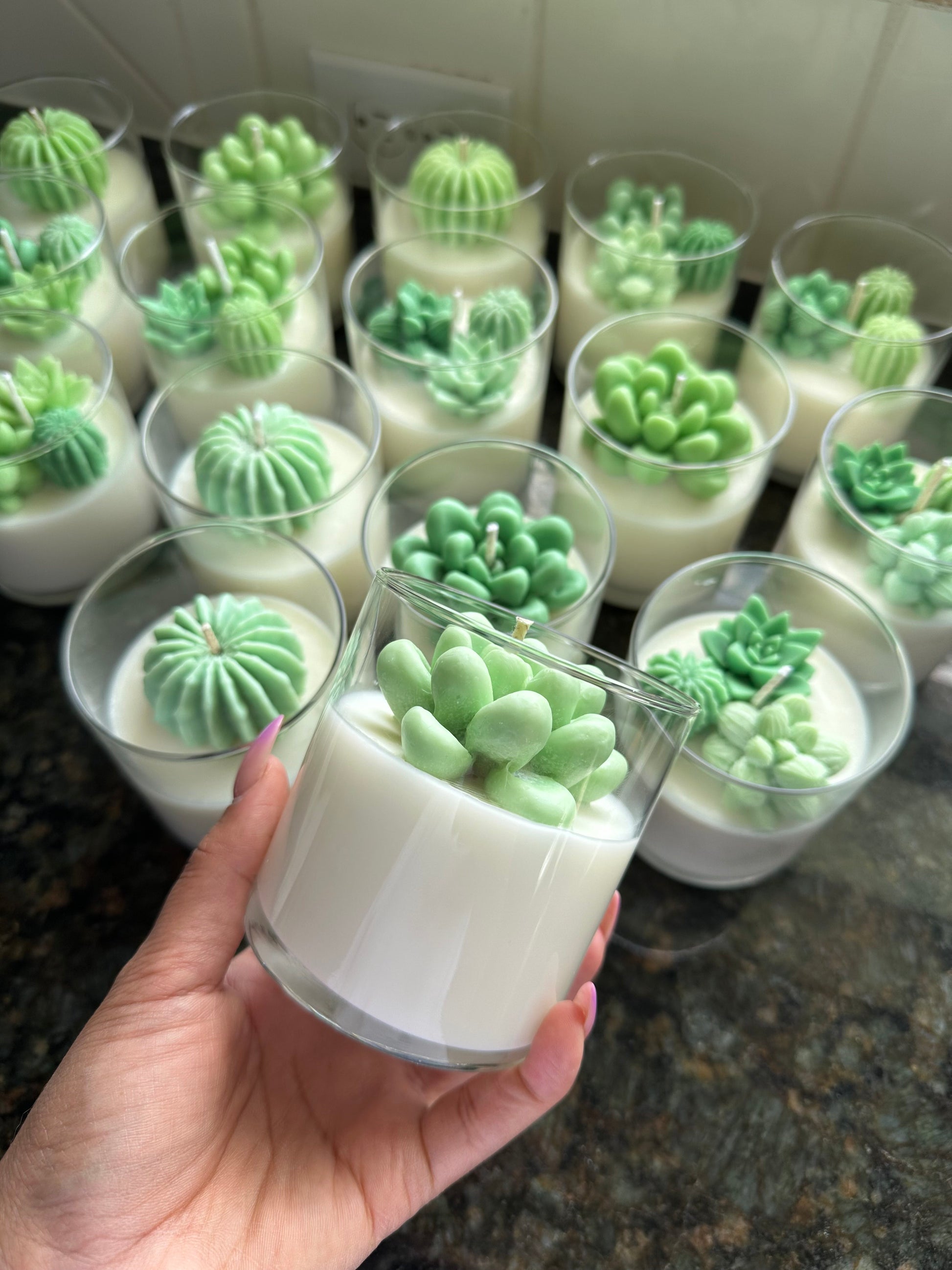 The image shows a collection of candles in clear glass containers, each topped with a highly detailed, green wax figure shaped like a succulent or cactus. The candles are creamy white, and the green succulents are placed at the top, making them the focal point. A hand is holding one of the candles in the foreground, displaying the intricate detail of the succulent design. The background features several other similar candles arranged closely together on a dark surface, likely a countertop.