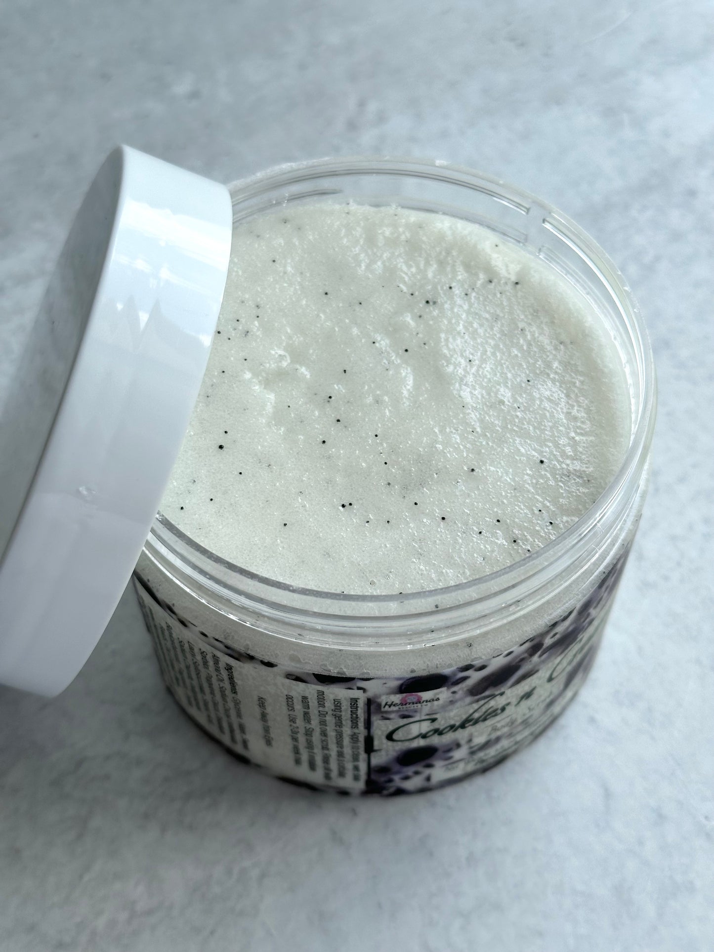 The image shows an open jar of "Cookies n Cream" body scrub from the brand "Hermanas Beauty Co." The scrub inside the jar has a creamy white texture with small black specks throughout, resembling cookie crumbs. The jar is clear, with the lid removed and placed beside it, revealing the scrub's smooth surface. The label on the jar features a "cookies and cream" theme, consistent with the appearance of the product. The background is a light, textured surface.