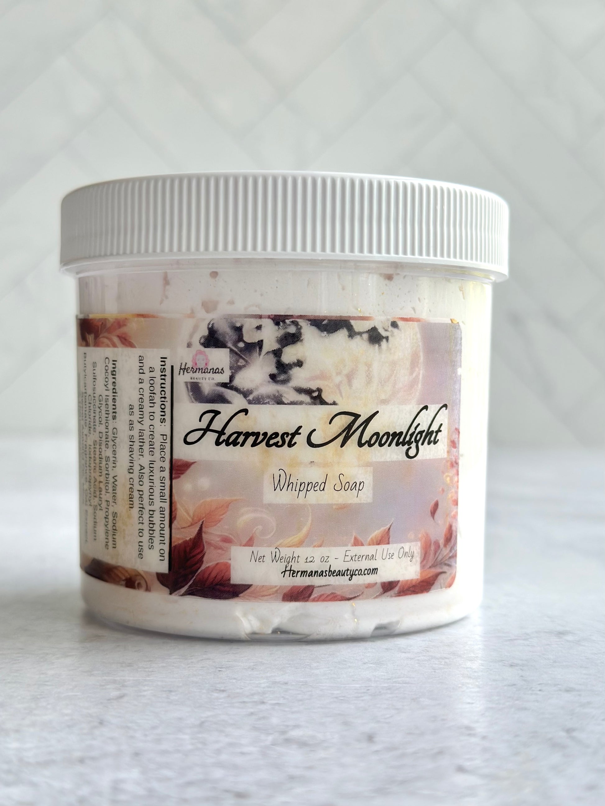 The image shows a jar of whipped soap labeled "Harvest Moonlight" from the brand "Hermanas Beauty Co." The jar is made of clear plastic with a white lid, allowing the creamy soap inside to be visible. The label features an autumn-themed design with leaves and warm, soft colors, complementing the product name. The whipped soap is in a 12 oz container and is intended for external use only. The brand's website is listed at the bottom of the label.