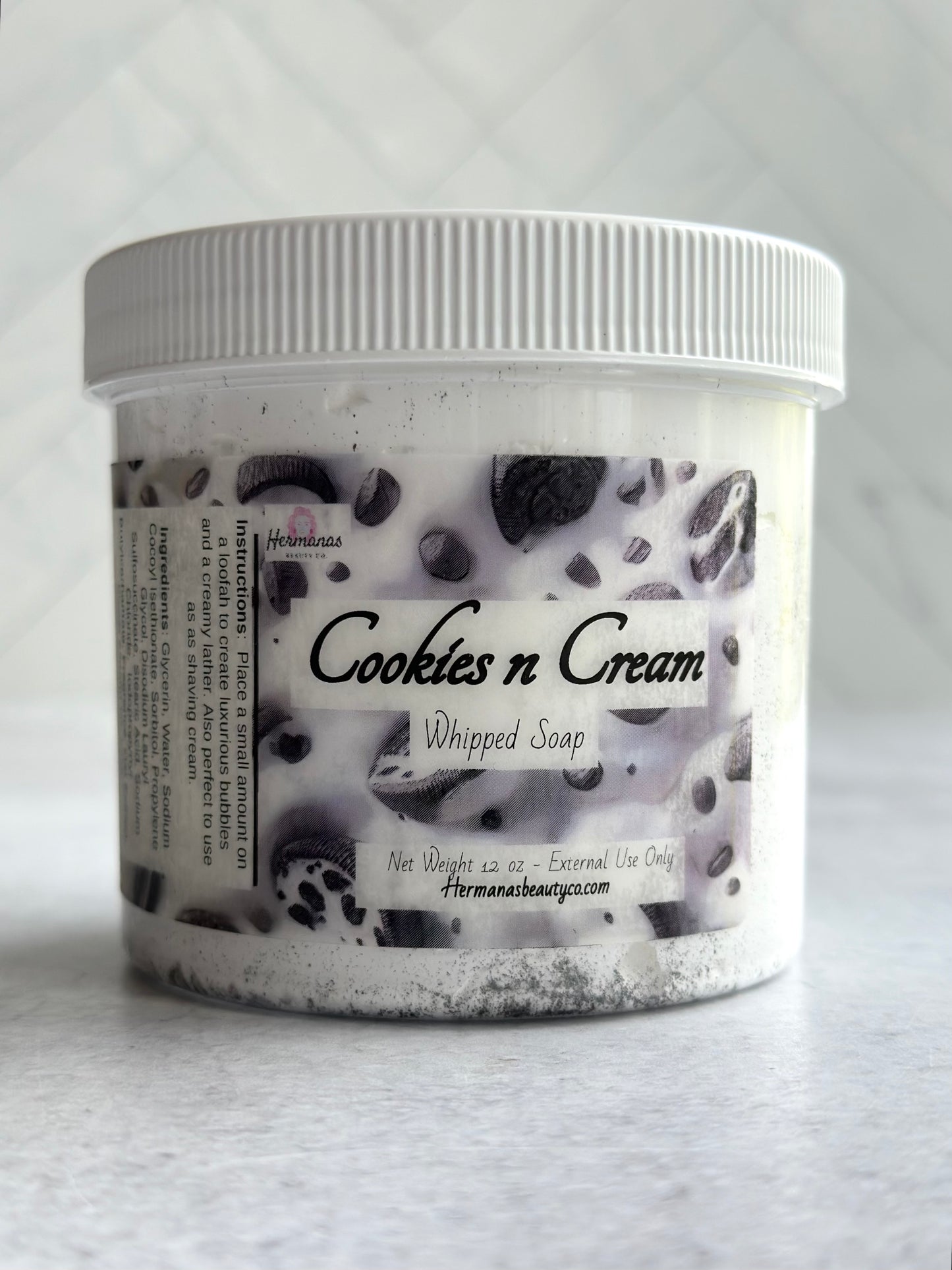 The image shows a jar of "Cookies n Cream" whipped soap from "Hermanas Beauty Co." The jar has a white lid and a label with a cookies and cream design, showcasing dark specks throughout a white base.