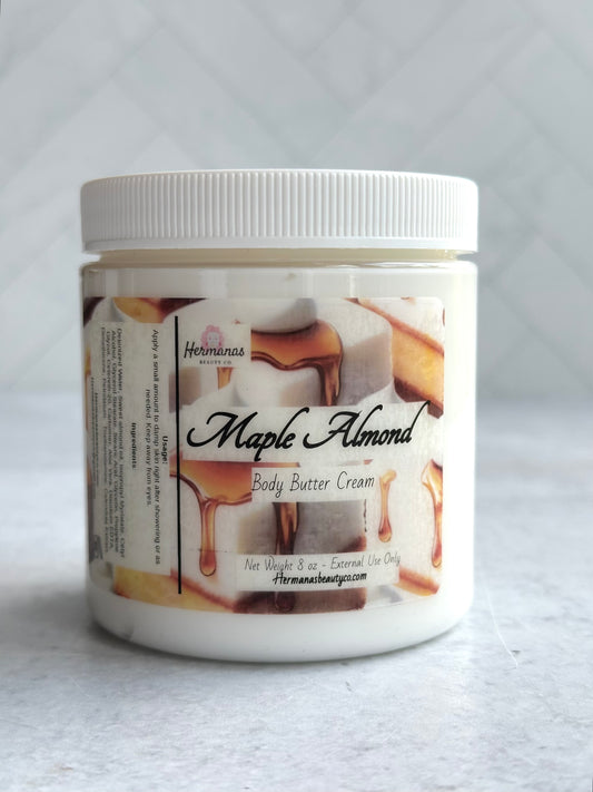 The image shows a white plastic jar of body butter cream labeled "Maple Almond" from the brand "Hermanas Beauty Co." The jar has a white lid and a label featuring images of maple syrup drizzling over what appears to be chunks of almond nougat or a similar confection. The product is described as "Body Butter Cream" with a net weight of 8 oz, intended for external use only. The label also includes the brand's website at the bottom.