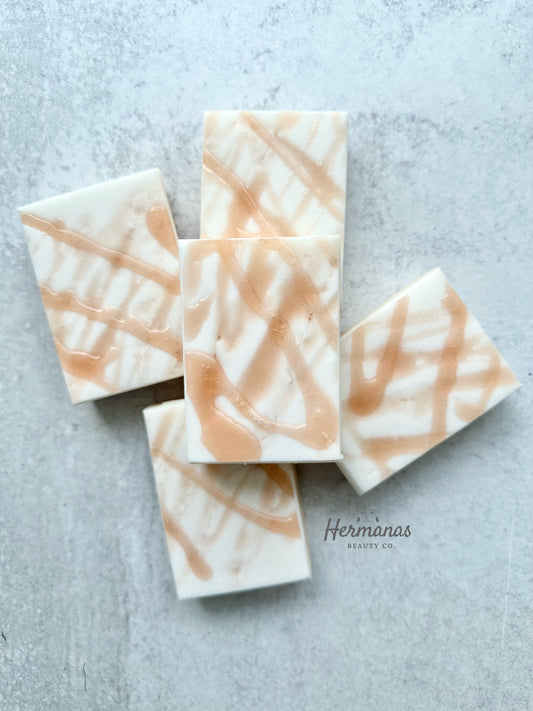 The image shows five rectangular soap bars arranged on a light-colored surface. The soaps have a creamy white base with swirling patterns of light brown or tan, giving them a marbled appearance. The swirling pattern is reminiscent of caramel drizzles or abstract brushstrokes. The brand name "Hermanas Beauty Co." is subtly displayed at the bottom of the image. The overall design of the soaps is elegant and visually appealing, with a natural and handcrafted aesthetic.