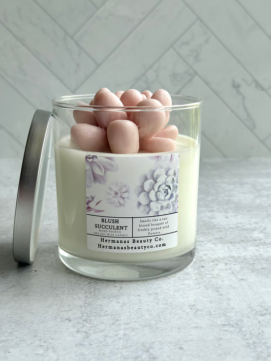 The image shows a glass jar candle labeled "Blush Succulent" from the brand "Hermanas Beauty Co." The candle is made of 100% soy wax and is topped with a detailed, pink wax figure shaped like a succulent plant. The label on the jar includes floral imagery and describes the scent as "smells like a sun-kissed bouquet of freshly picked wildflowers." The candle has a silver lid, which is placed leaning against the jar. The background features a light, marble-tiled wall.