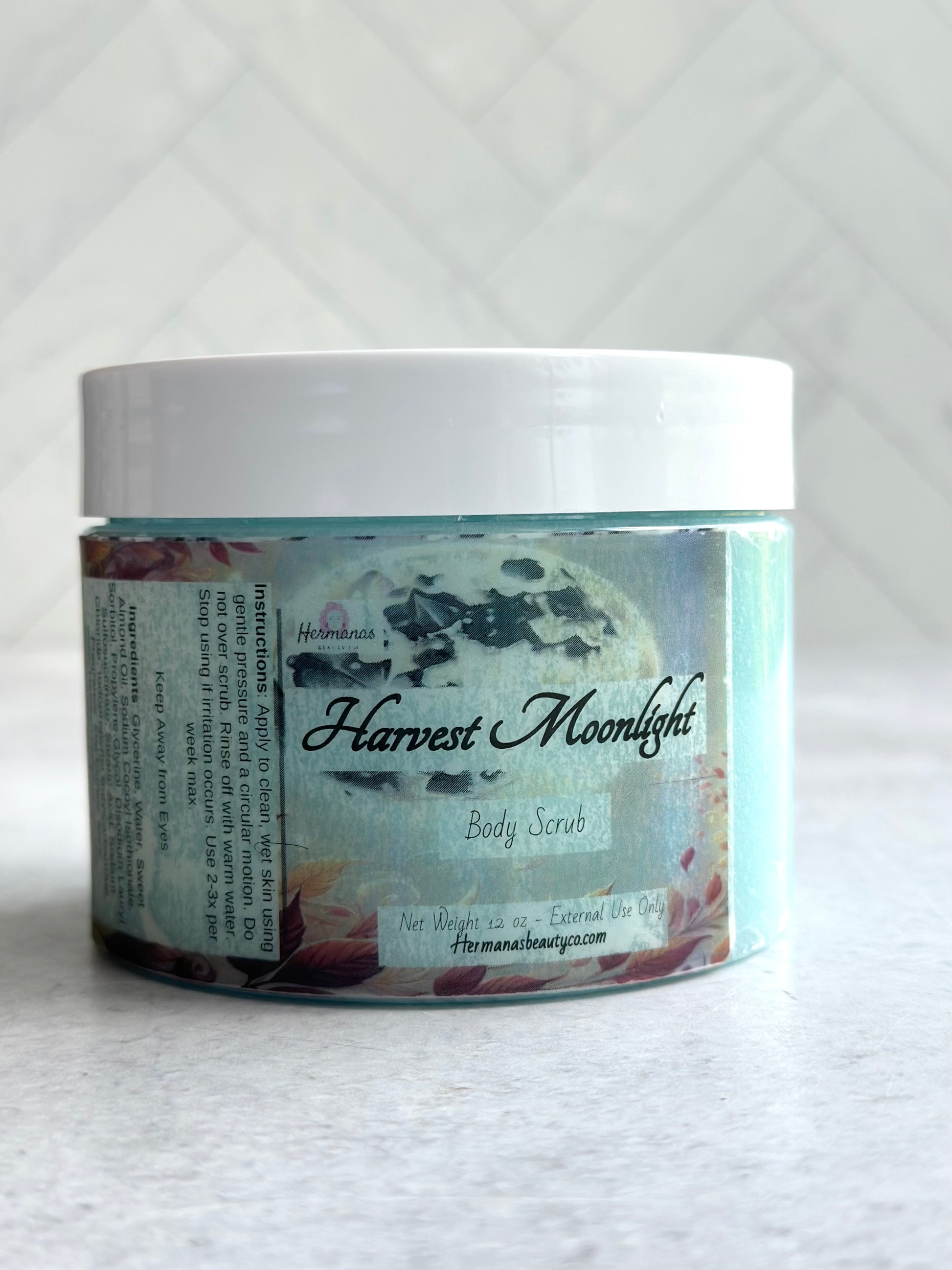 The image shows a jar of "Harvest Moonlight" body scrub from "Hermanas Beauty Co." The jar is clear with a white lid, revealing the light blue scrub inside. The label features a moonlit, autumn-themed design.