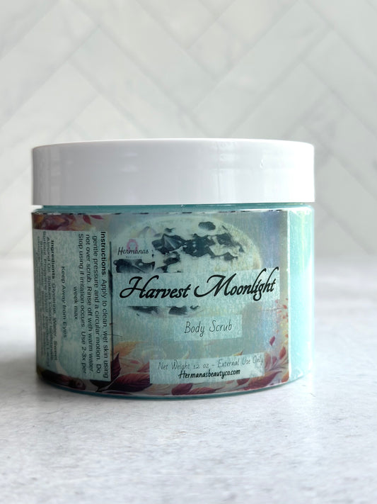 The image shows a jar of "Harvest Moonlight" body scrub from "Hermanas Beauty Co." The jar is clear with a white lid, revealing the light blue scrub inside. The label features a moonlit, autumn-themed design.