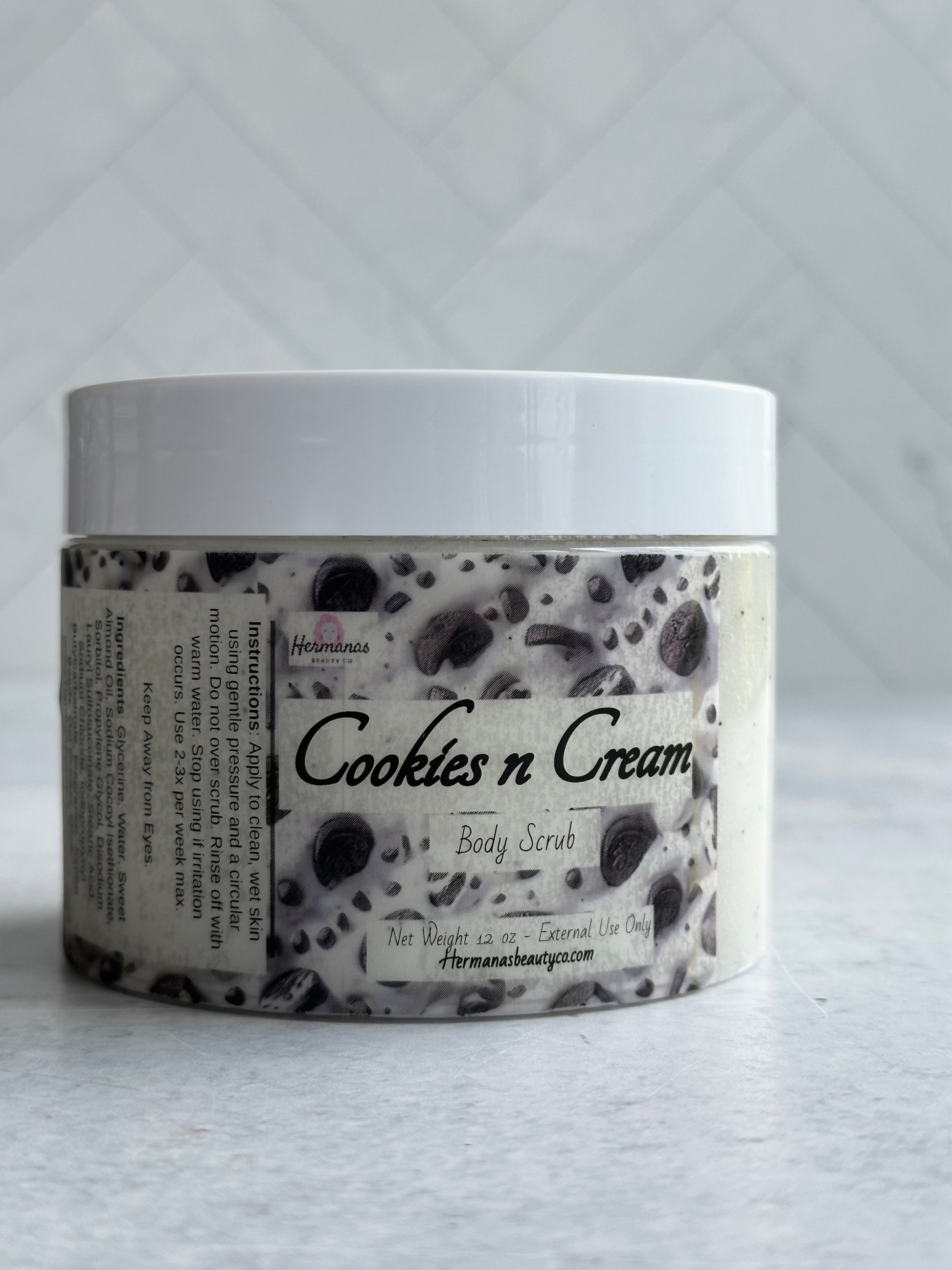 The image shows a jar of body scrub labeled "Cookies n Cream" from the brand "Hermanas Beauty Co." The jar is made of clear plastic with a white lid, revealing the scrub inside, which has a white base with dark specks resembling cookie crumbs. The label features an image that mimics the appearance of cookies and cream, enhancing the theme of the product. The scrub is packaged in a 12 oz container, and the label indicates it is for external use only. The brand's website is also listed on the label.