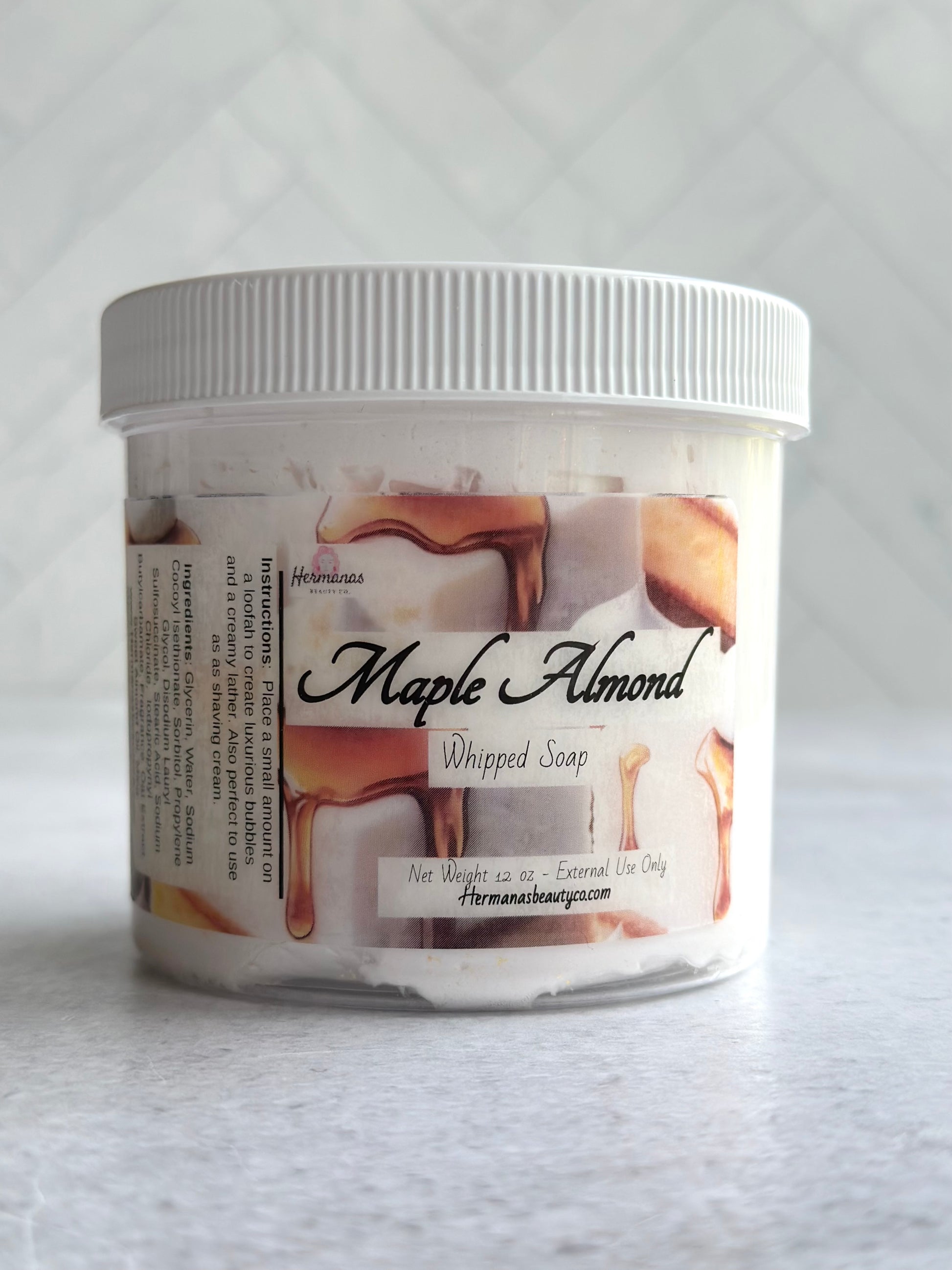 The image displays a jar of whipped soap labeled "Maple Almond" from the brand "Hermanas Beauty Co." The jar is made of clear plastic with a white lid, revealing the whipped soap inside. The label features images of maple syrup drizzling over almond cake pieces, reflecting the product's scent theme. The whipped soap is packaged in a 12 oz container, and the label indicates it is for external use only. The brand's website is also listed on the label.