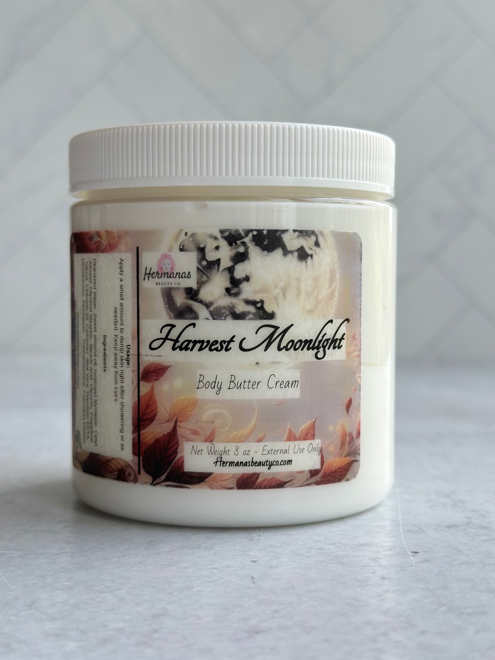 The image shows a jar of "Harvest Moonlight" body butter cream from "Hermanas Beauty Co." The jar is white with a white lid and features an autumn-themed label with leaves and a moonlit design.