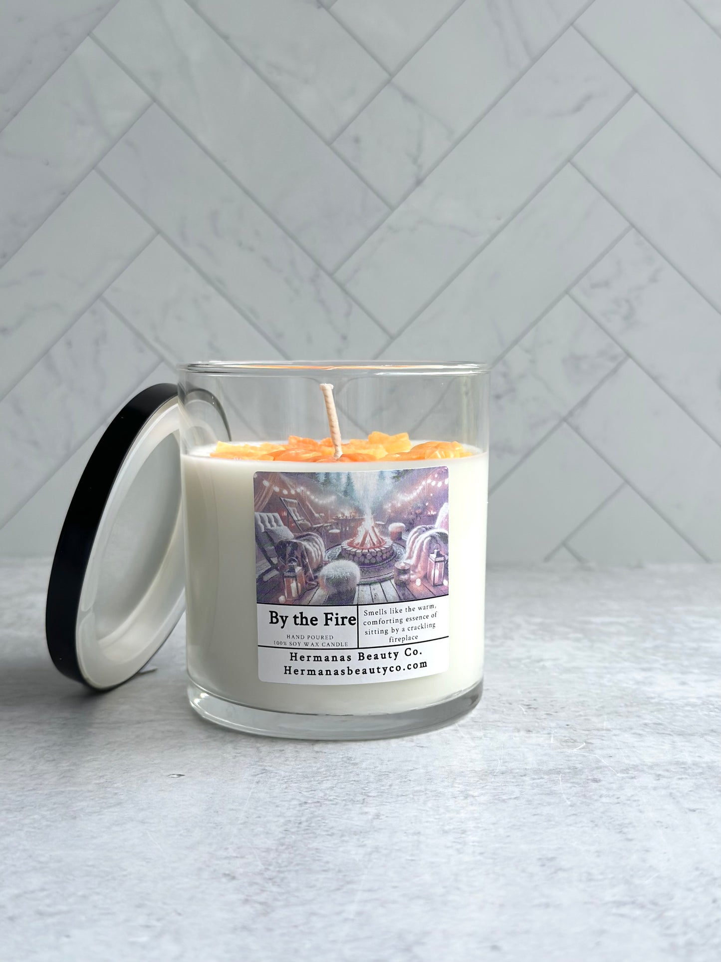 The image shows a glass jar candle labeled "By the Fire" from the brand "Hermanas Beauty Co." The candle is white and topped with a wax design that mimics a glowing fire with warm orange and yellow tones. The label features an illustration of a cozy outdoor fire pit surrounded by chairs, with the description "Smells like the warm, comforting essence of sitting by a crackling fireplace." The candle has a black lid, which is placed leaning against the jar.