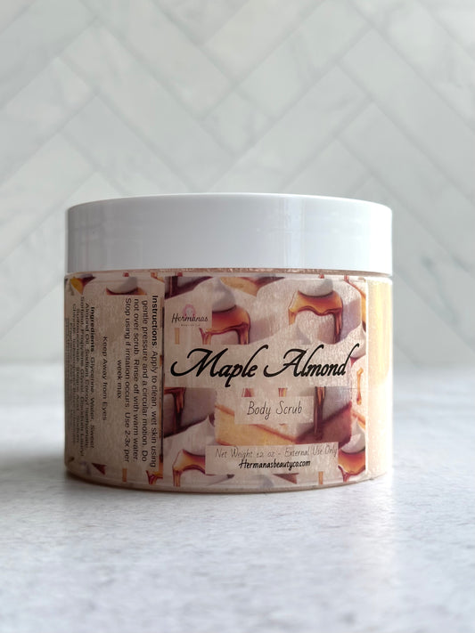 The image displays a jar of body scrub labeled "Maple Almond" from the brand "Hermanas Beauty Co." The jar is made of clear plastic with a white lid, allowing the scrub inside to be slightly visible. The label features images of maple syrup drizzling over almond cake pieces, emphasizing the product's scent theme. The body scrub is packaged in a 12 oz container, and the label indicates it is for external use only. The brand's website is also mentioned on the label.