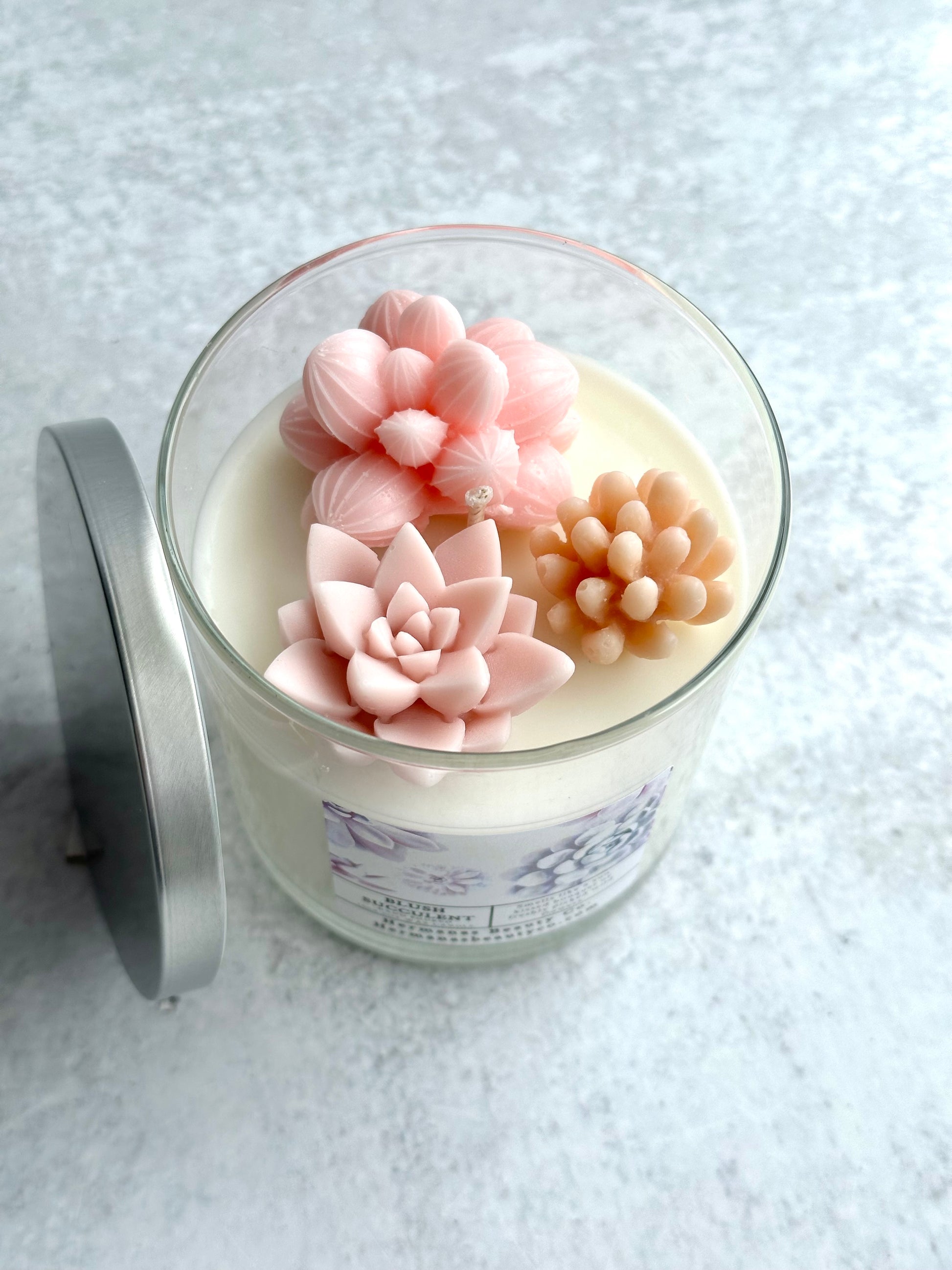 The image shows a glass jar candle with a creamy white wax base, topped with three intricately designed wax succulents in soft pink and beige tones. The candle has a silver lid placed beside the jar. The succulents are detailed and realistic, adding a decorative touch to the candle. The label on the jar reads "Blush Succulent" and includes floral designs, consistent with the overall delicate and elegant aesthetic of the product. The candle is set on a light, textured surface, creating a clean look.