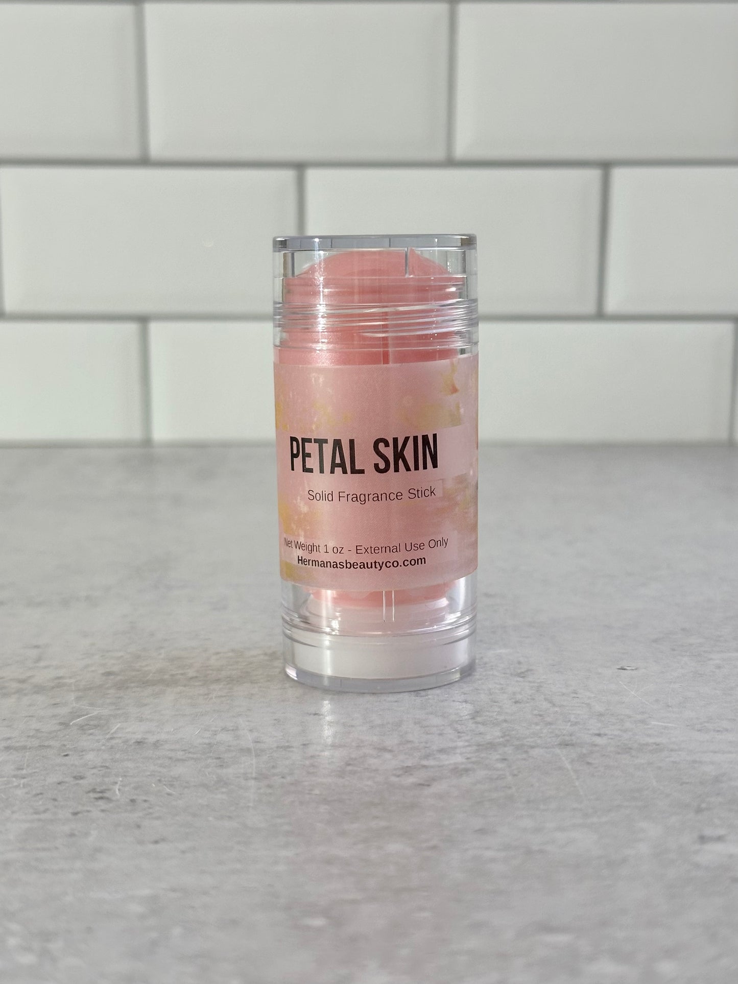 Solid fragrance stick labeled 'Petal Skin' on a gray surface with a tiled wall background.