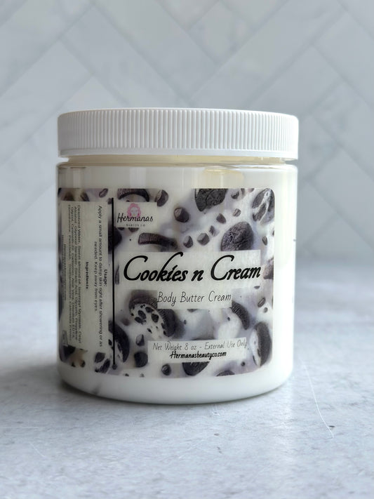 The image shows a jar of "Cookies n Cream" body butter cream from "Hermanas Beauty Co." The jar has a white lid and a label with a cookies and cream theme, featuring images of cookies and dark specks throughout the design.