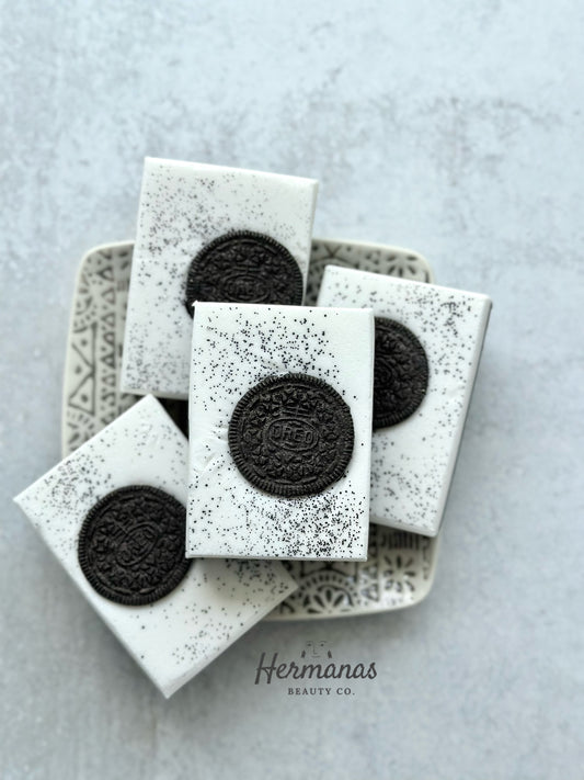 The image displays four rectangular white soaps arranged on a decorative plate. Each soap has a realistic, embedded black cookie in the center, with a fine sprinkle of what appears to be black specks, resembling cookie crumbs, scattered over the top surface. The brand name "Hermanas Beauty Co." is visible at the bottom of the image.