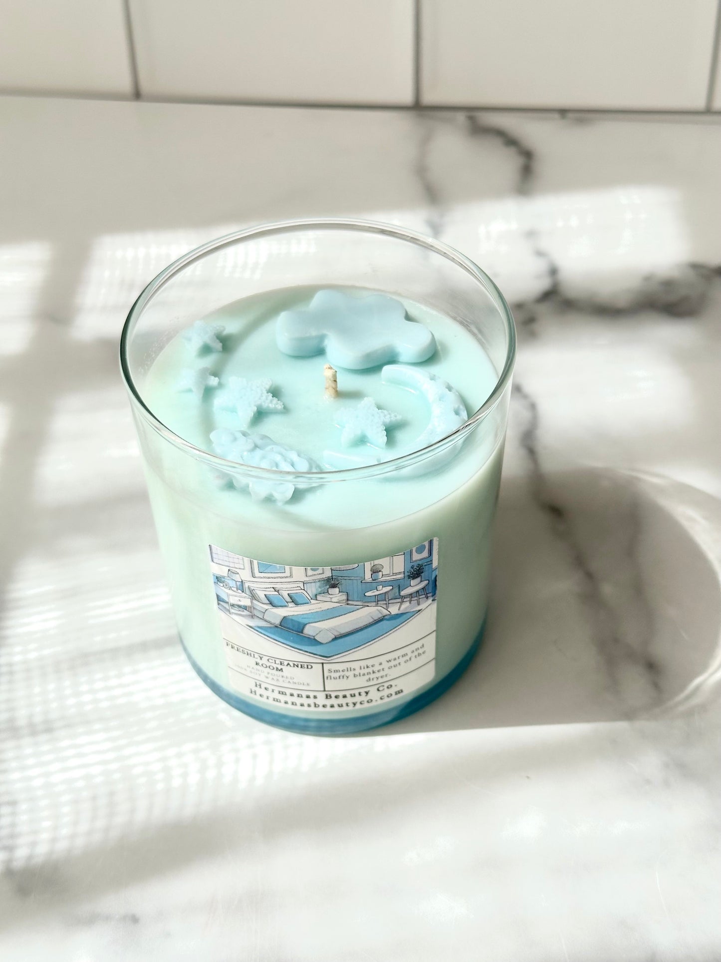 Light blue candle in clear glass jar, has a white and blue label and clouds, moons and stars as a detail on the top.