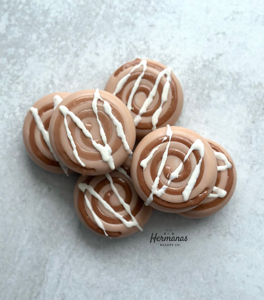 The image shows a group of round, swirl-patterned soaps designed to resemble cinnamon rolls. The soaps are a light brown color with darker brown swirls, and they are topped with a white drizzle that mimics icing. The soaps are stacked together on a light, textured surface, with the brand name "Hermanas Beauty Co." visible at the bottom of the image. The overall appearance is highly realistic, evoking the look of freshly baked cinnamon rolls.