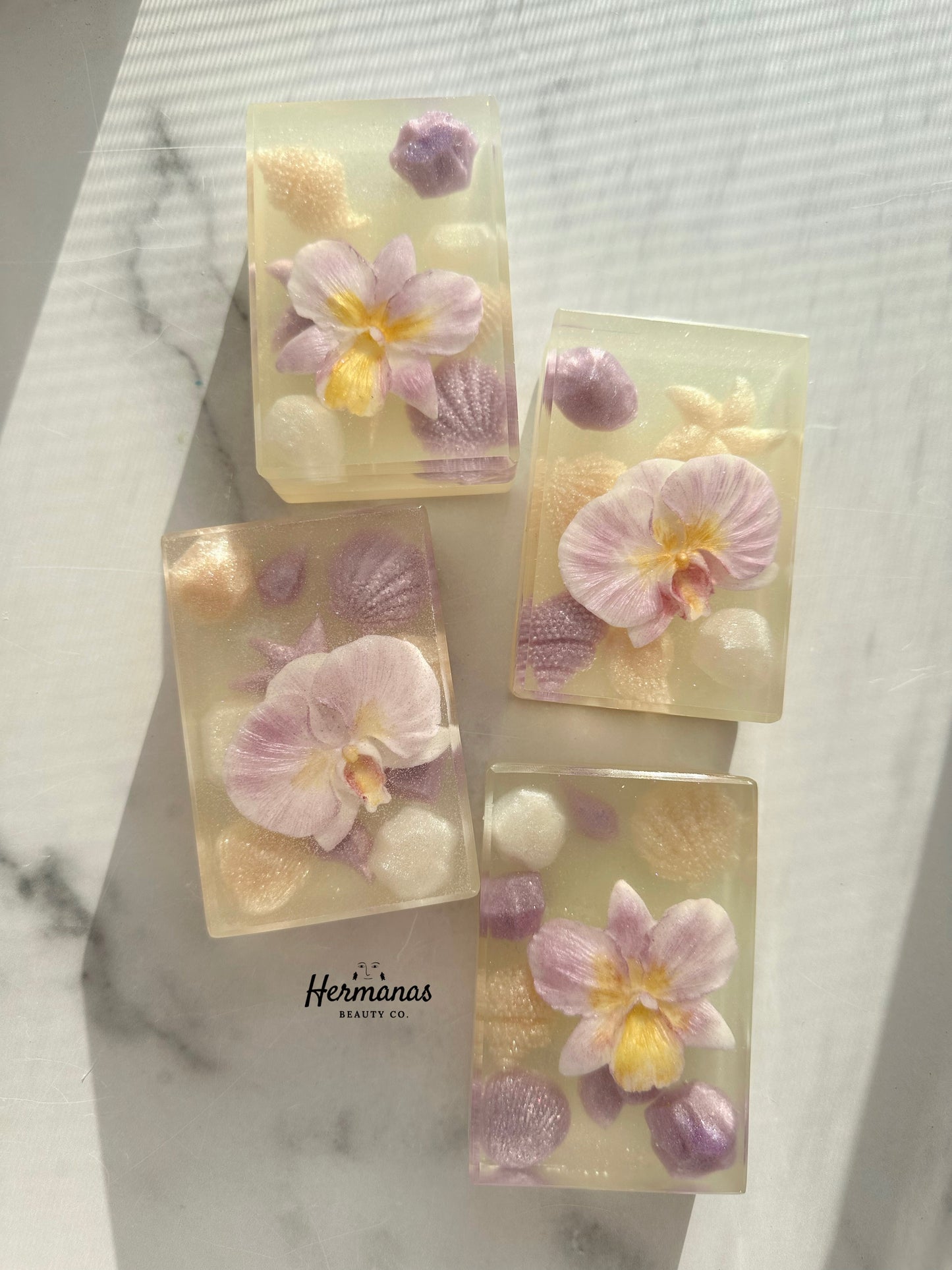 Decorative soap bars with pink flowers and seashells on a marble surface, featuring the brand 'Hermanas Hermanas'.
