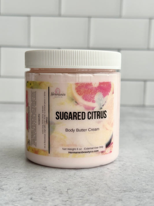 Jar of 'Sugared Citrus' body butter cream on a gray surface with a tiled wall background.