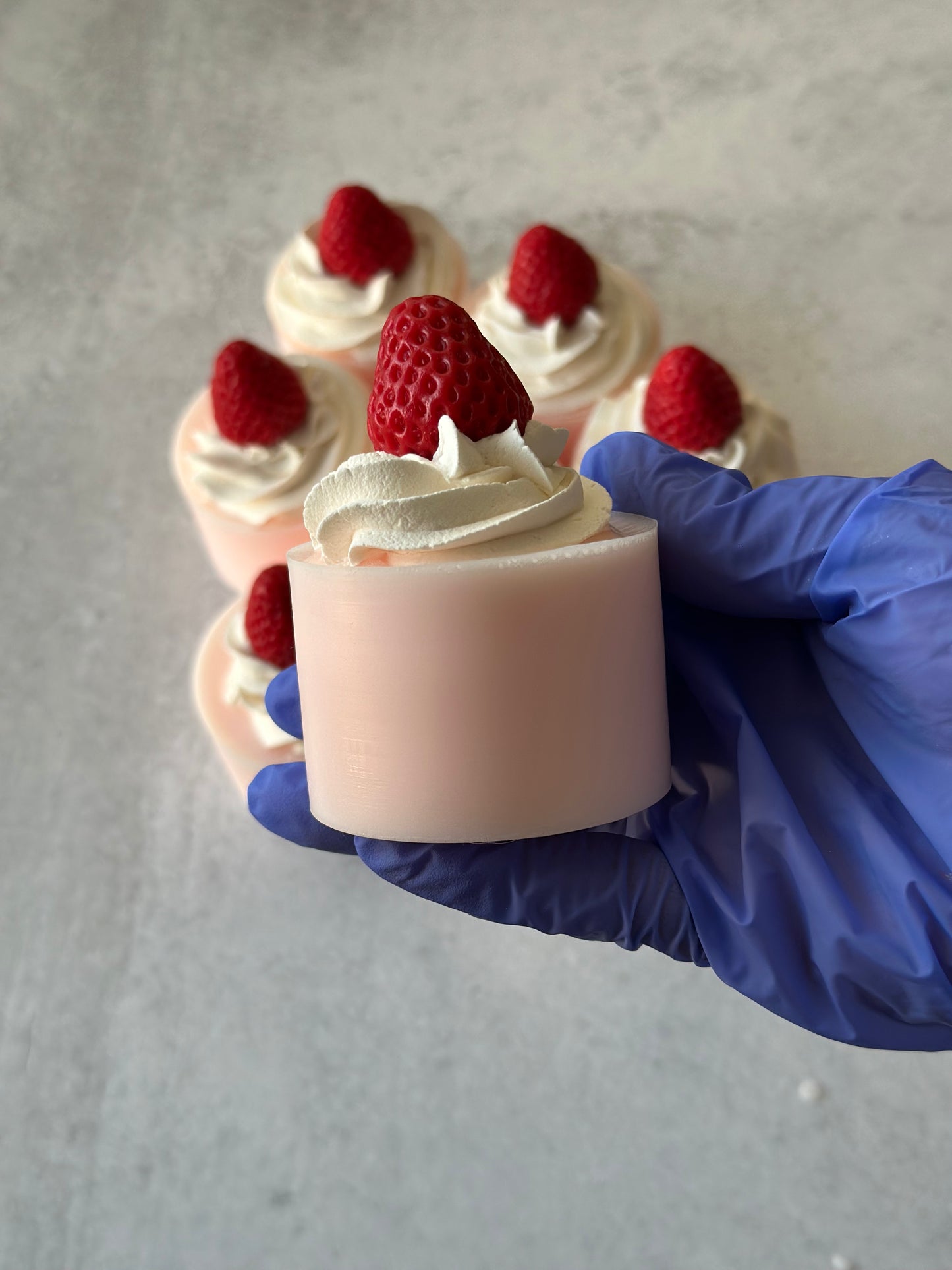 Hand holding a small dessert soap with whipped cream and a strawberry, with more dessert soaps in the background.