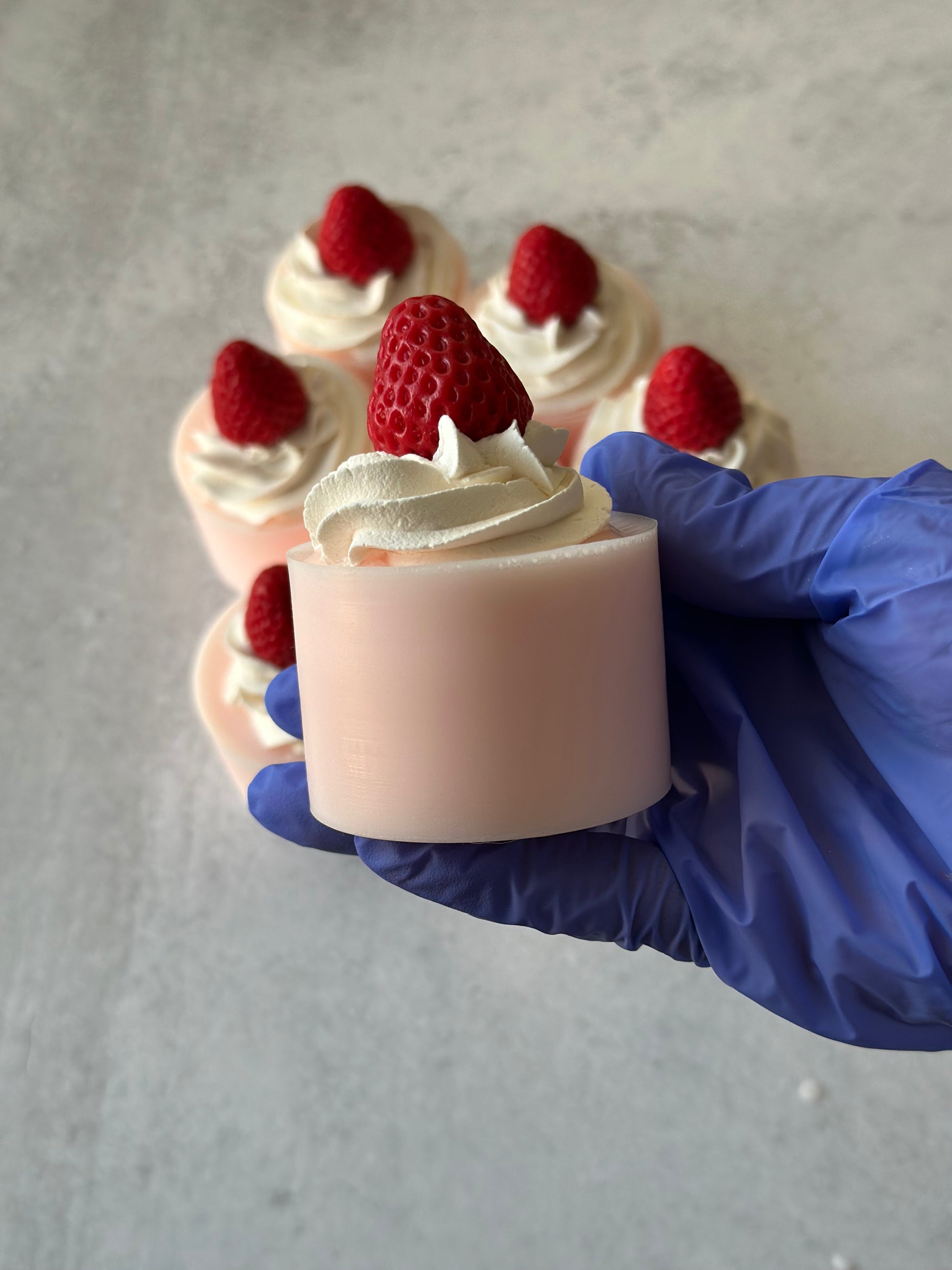 Hand holding a small dessert soap with whipped cream and a strawberry, with more dessert soaps in the background.