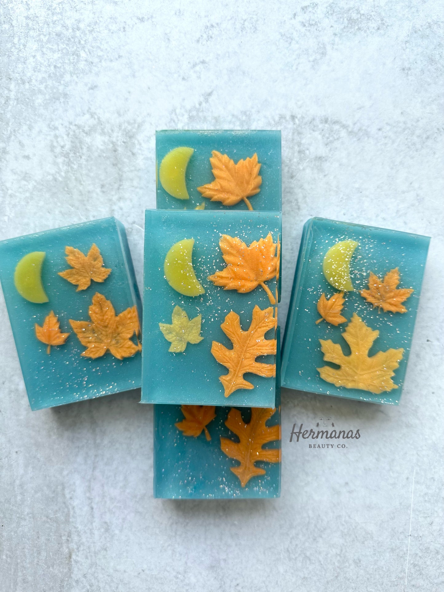 The image shows five beautifully crafted blue soaps adorned with autumn-themed decorations. Each soap features detailed yellow and orange leaves, along with a crescent moon shape, all set against a blue background. The leaves and moons are embedded in the soap, with a subtle sparkle or shimmer visible across the surface. The soap is labeled with the brand "Hermanas Beauty Co." in the corner.