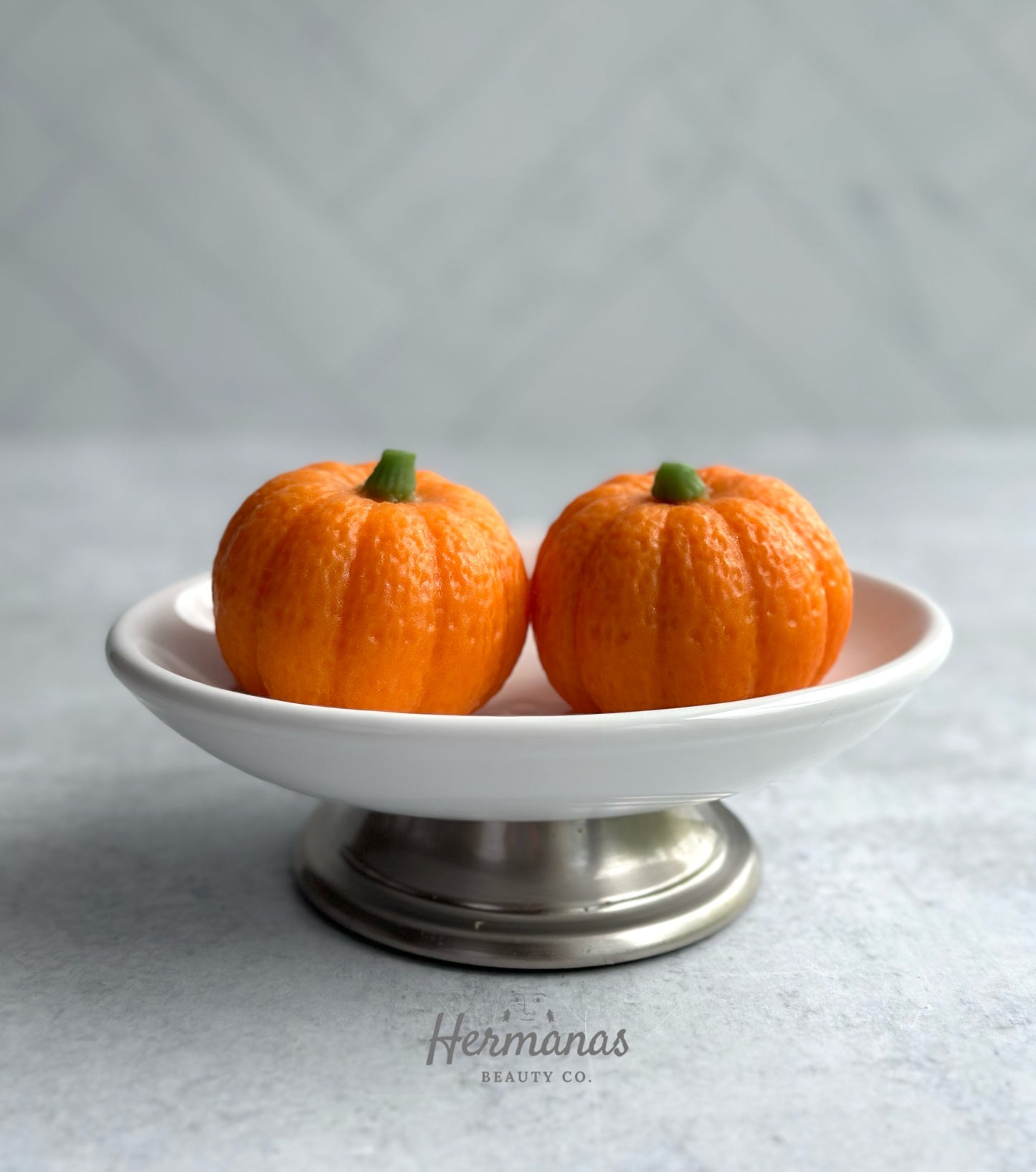 The image shows two small, orange pumpkin-shaped soaps placed on a white pedestal dish with a silver base. The pumpkins are detailed with a textured surface to resemble real pumpkins, and they each have a small green stem on top. The dish is set against a light, neutral background, and the brand name "Hermanas Beauty Co." is visible at the bottom of the image. The overall presentation is simple yet festive, evoking a fall or Halloween theme.