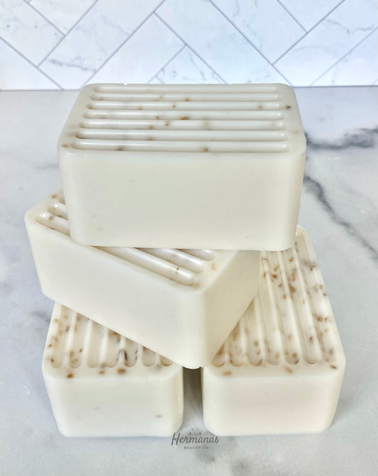 The image shows a stack of four rectangular soap bars, each with a ridged surface on one side. The soaps are a creamy off-white color with small specks, likely exfoliating ingredients, visible throughout the bars. The soaps are neatly arranged on a smooth, light-colored surface, with the brand name "Hermanas Beauty Co." subtly displayed at the bottom of the image. The background features a white, marble-like tile wall with a geometric pattern.