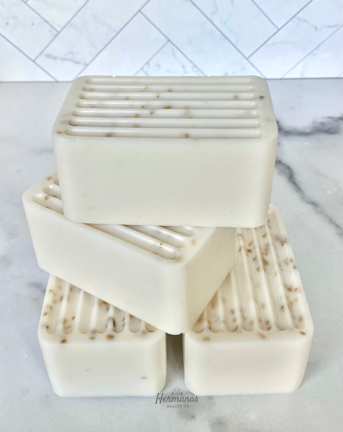 The image shows a stack of four rectangular soap bars, each with a ridged surface on one side. The soaps are a creamy off-white color with small specks, likely exfoliating ingredients, visible throughout the bars. The soaps are neatly arranged on a smooth, light-colored surface, with the brand name "Hermanas Beauty Co." subtly displayed at the bottom of the image. The background features a white, marble-like tile wall with a geometric pattern.